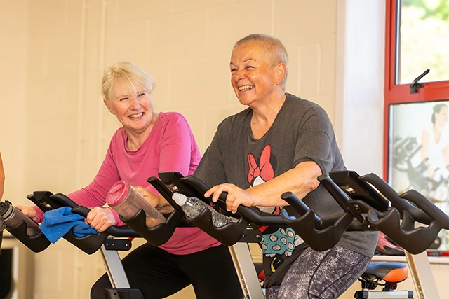 Revolution Two women smile whilst riding indoor bicycles as part of a Revolution class. Both wear gymwear and have water bottles.