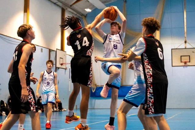 Basketball A group of young people play basketball ina sports hall. Two players in the middle have jumped. One tries to stop the opther from shooting a basket.