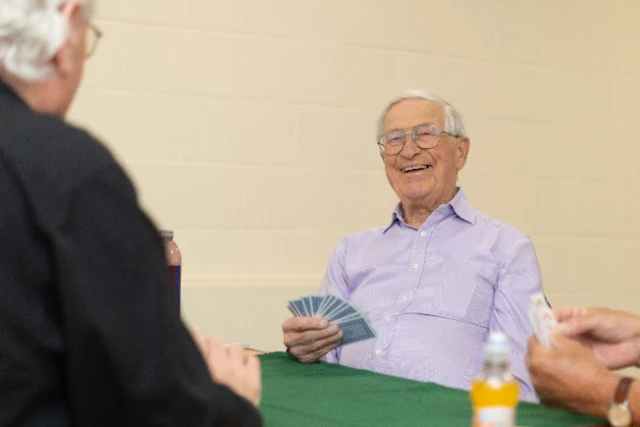 Three older players take part in the card game Bridge, each holding playing cards in their hands, sitting around a table covered in a green cloth.