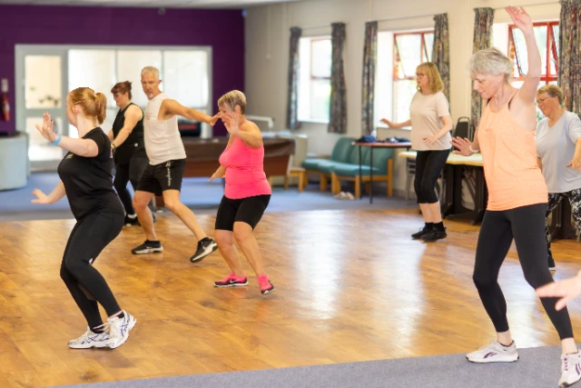 Dance Fit (8) An instructor leads a class of Dance Fit. Attendees in colourful gym wear reach upward mid-move.