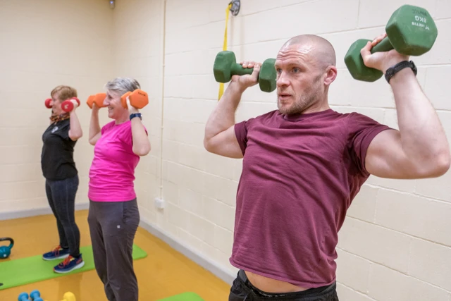 Functional Strength (12) Three people are exercising with dumbbells in a gym. A man in the foreground is performing an overhead press with green dumbbells, while two women in the background are doing the same exercise with orange and red dumbbells respectively.