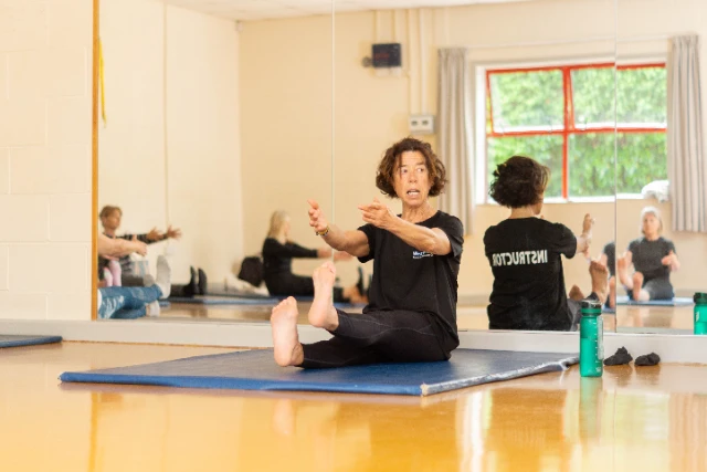 Tone and Stretch (46) Female class leader demonstrates a move within the tone and stretch class. Attendees can be seen replicating this move in the mirror.