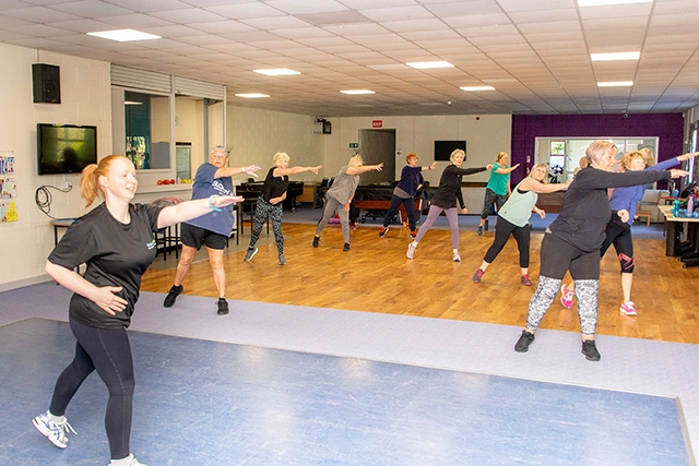 Bodyfit (2) A group of women, led by an instructor in the foreground, are participating in an exercise class in a community hall. They are all in workout attire and performing similar arm movements.