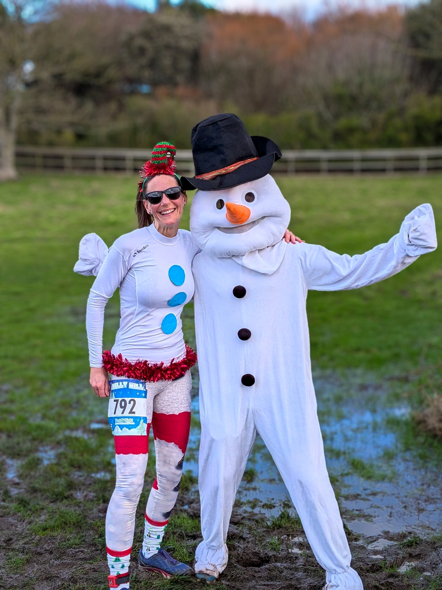 Chilly Hilly Mascot A woman dressed in a festive snowman costume poses with a person in a full snowman mascot suit. They are standing outdoors in a grassy area with trees in the background.