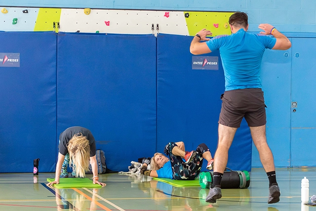 Circuit Training (46) An instructor in a blue shirt and shorts stands facing away from the camera, leading a circuits class. Two women are on the floor on mats, one in a plank position and the other lying on her back.