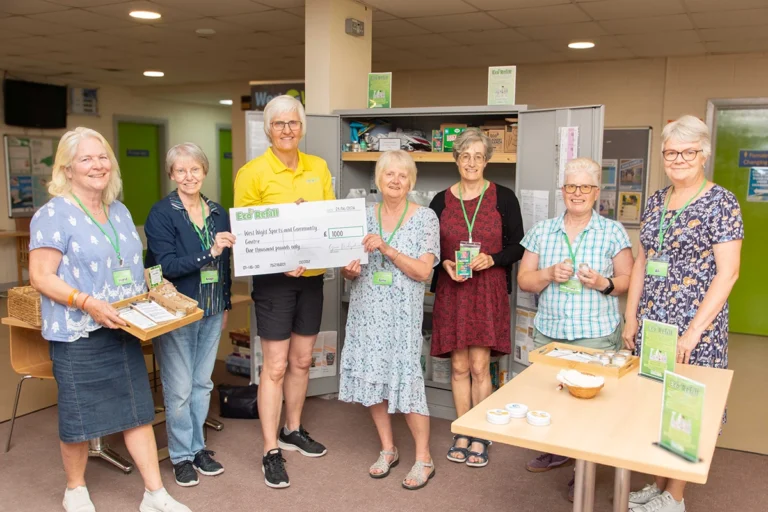 A group of female Eco Refill volunteers are posing for a photo, alongside centre manager, Clare Griffin. They are presenting a £1000 cheque. Several women are wearing lanyards with name tags. There are also items displayed on a table in the foreground, related to the eco-refill service.