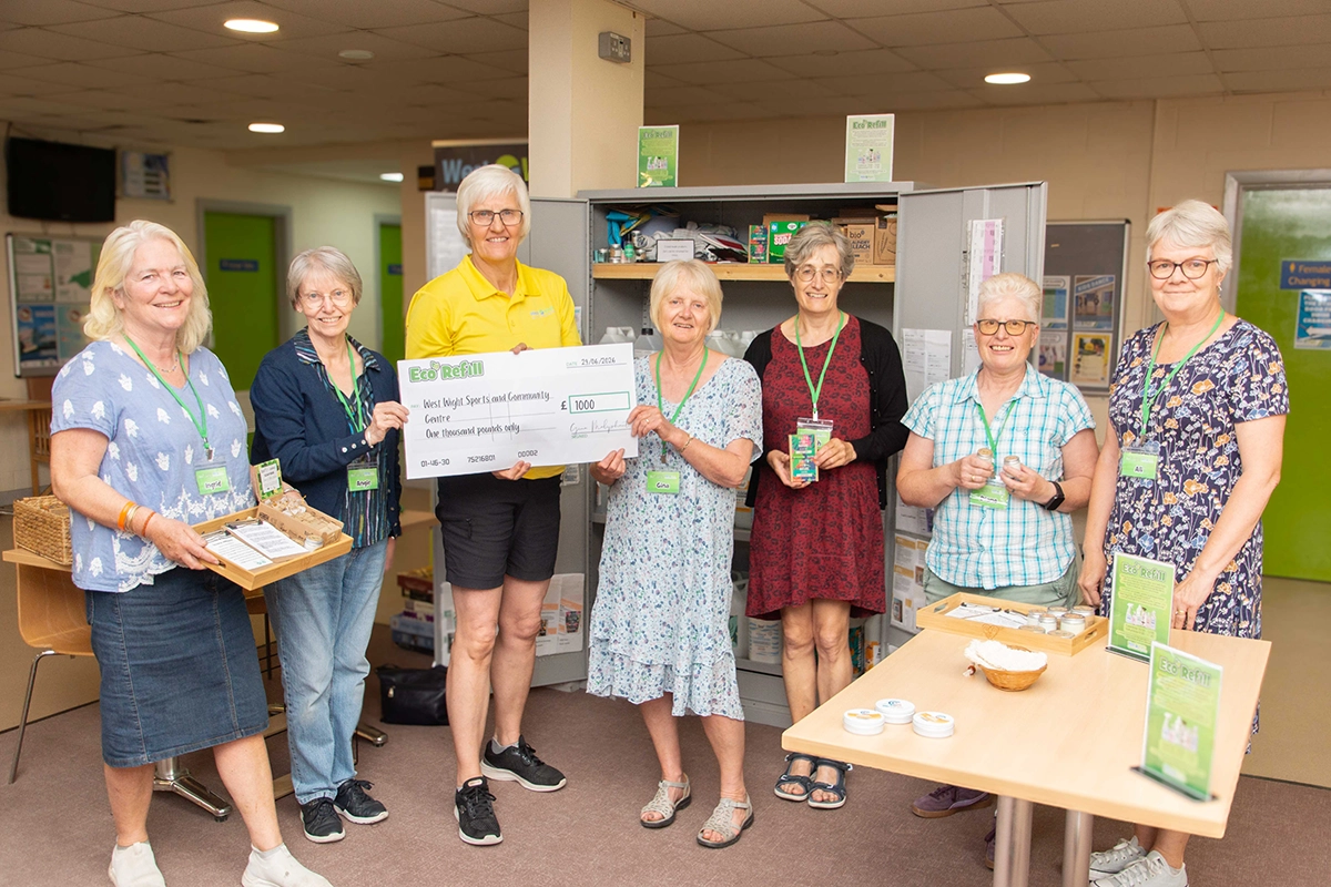 Eco Refill 29-06-2026 copy A group of female Eco Refill volunteers are posing for a photo, alongside centre manager, Clare Griffin. They are presenting a £1000 cheque. Several women are wearing lanyards with name tags. There are also items displayed on a table in the foreground, related to the eco-refill service.