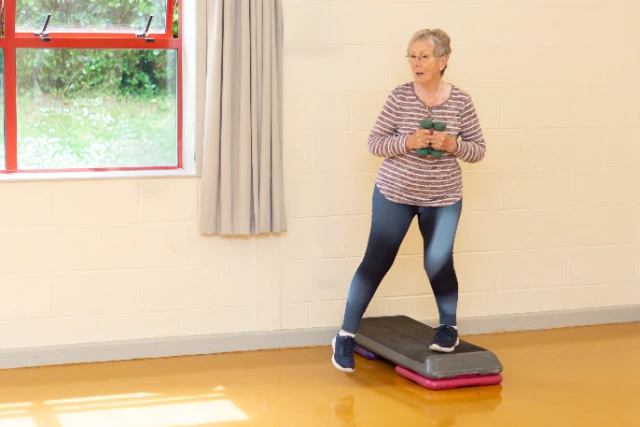Gentle Circuits (45) An elderly woman is exercising with hand weights while standing on a step aerobics platform. She is wearing a striped long-sleeved shirt and ombre leggings.