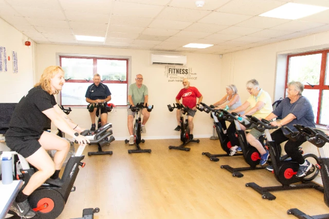Gentle Revolution A group of older adults is participating in a Gentle Revolution cycling class led by a younger instructor. The class is taking place in a bright, indoor fitness room.