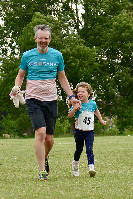 A man holding a teddy bear and a young girl are running together on a grassy field. They are both wearing matching shirts.