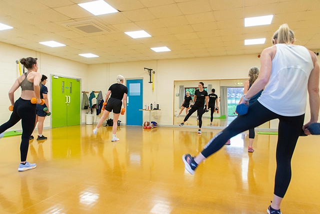 Legs Bums and Tums A group of women are participating in a fitness class, performing exercises with dumbbells. They are in a brightly lit room with a shiny yellow floor and mirrors on one wall.