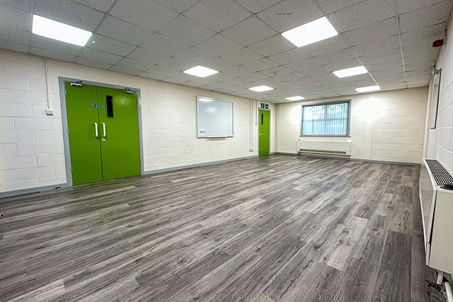 Meeting Room This is an interior view of an empty meeting room with grey wood-look flooring. The room features white brick walls, fluorescent lighting, and two bright green doors.