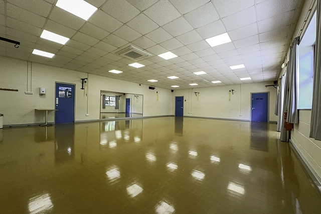 Mountbatten Room An empty dance studio with a polished floor and mirrors on the wall. There are two blue doors visible and large windows with curtains.