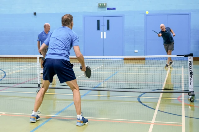 Pickleball (2) Three men play pickleball indoors on a court. One man is hitting the ball, while two others are positioned on the other side of the net.