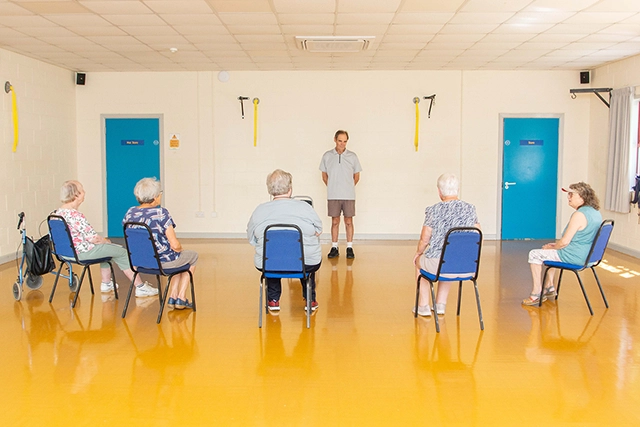 A group of older adults are seated in chairs facing a male instructor in a room with a bright yellow floor. The instructor stands in front of them, and they appear to be participating in the Reall Useful Cafe session.