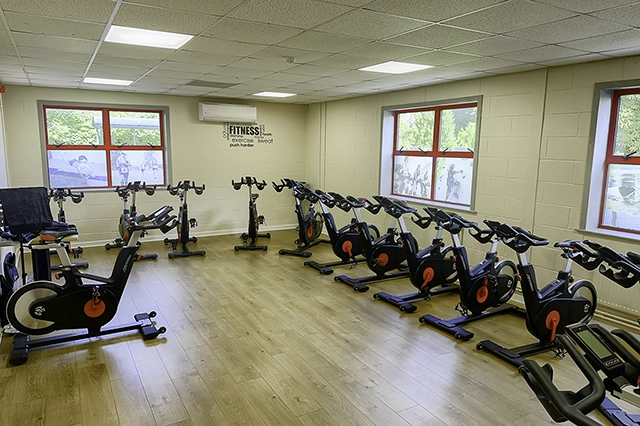 Revolution studio An indoor cycling studio is filled with rows of black and red stationary bikes. The room has a light-colored wooden floor and beige walls with windows showing outdoor scenery and motivational text.