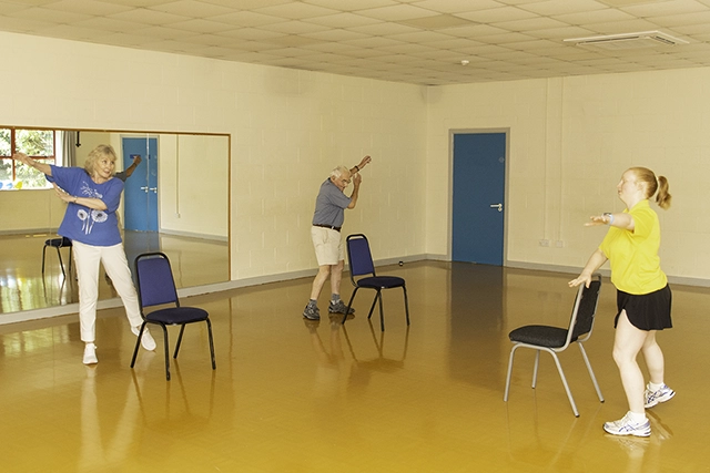 Strength and Balance (1) Two people are participating in a Strength and Balance exercise class in a room with a mirrored wall. They are all in motion, with one woman in blue and an older man in a blue shirt and shorts, led by a female instructor in a yellow shirt.