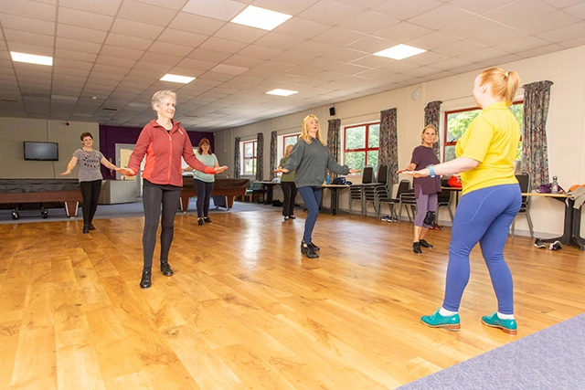 Tap A group of women are participating in a tap dance class in the community hall. They are all standing and following along with an instructor who is facing away from the camera.