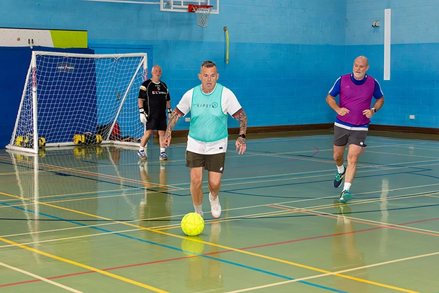 Veterans Football Three older men are playing Veterans Football in the sports hall. One man is dribbling a bright yellow soccer ball while two others are running on the court.